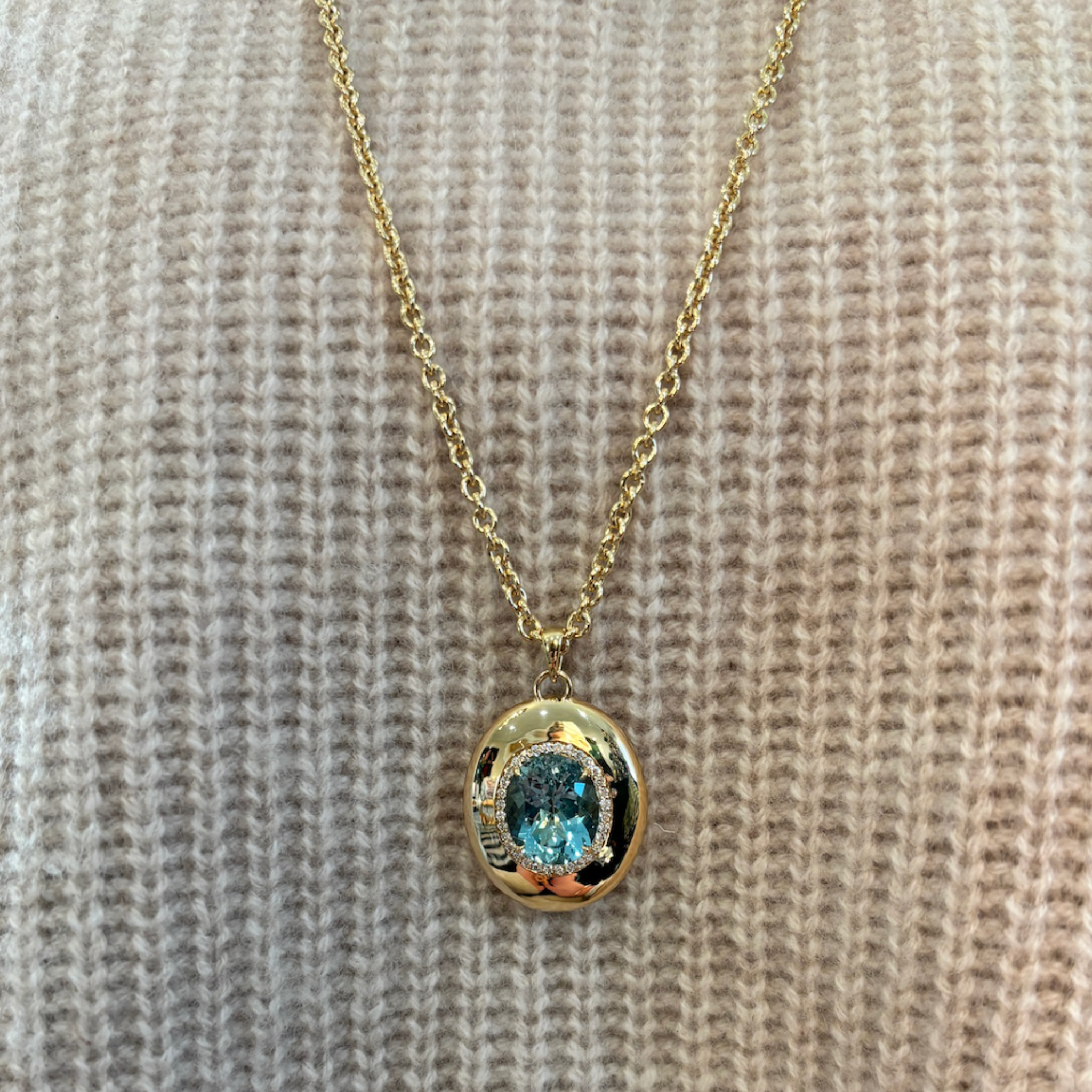 The Pavé Oval Cut Aquamarine Collet Pendant in Yellow Gold is worn over a cream-colored knitted sweater.