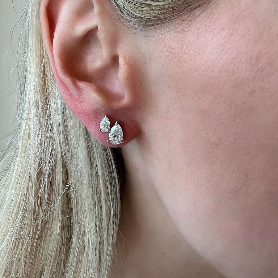 A woman with fair skin and straight, light blonde hair wears the Duo Studs in White Gold with Pear Diamonds—featuring one larger and one smaller pear diamond for an elegant, made-to-order look.