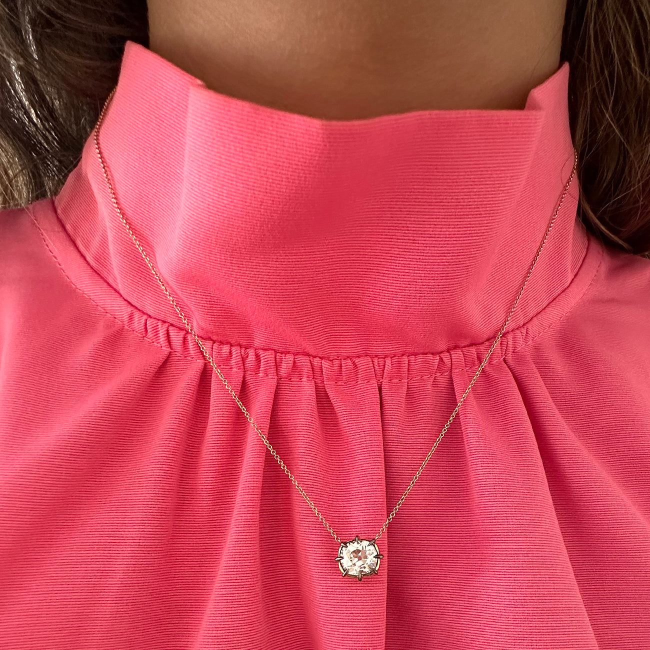 A close-up of a person wearing a vibrant pink high-neck blouse and the Cushion Cut Diamond Collet Pendant in Yellow and Blackened Gold, featuring a delicate flower-shaped design with a central cushion cut diamond.