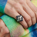 A close-up of a hand resting on another, featuring the Baguette and Marquise Sapphire Engagement Ring Jacket in white gold with dazzling sapphires and a large diamond. The person wears a colorful, striped fuzzy sweater.