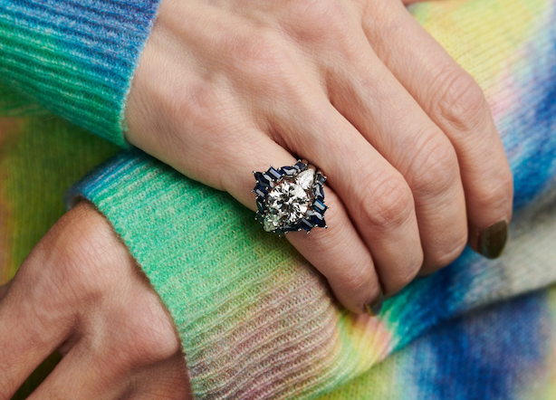 A close-up of hands wearing the Bespoke Baguette and Marquise Sapphire Engagement Ring Jacket in White Gold, featuring shimmering sapphires and blue accents, paired with a vibrant tie-dye sweater in green, blue, and yellow.