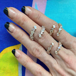 A hand with a geometric black and clear French manicure displays six 18K gold rings, including the Wraparound Ring with Mixed Shape Diamonds, against a colorful abstract background.