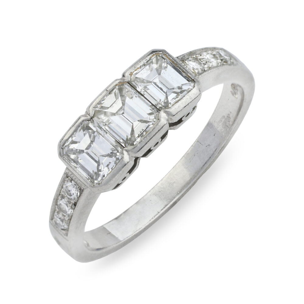 This Three-Stone Diamond Ring, inspired by a 1950 vintage style, showcases three central emerald-cut diamonds flanked by round diamonds on each side and features classic platinum hallmarks for authenticity.