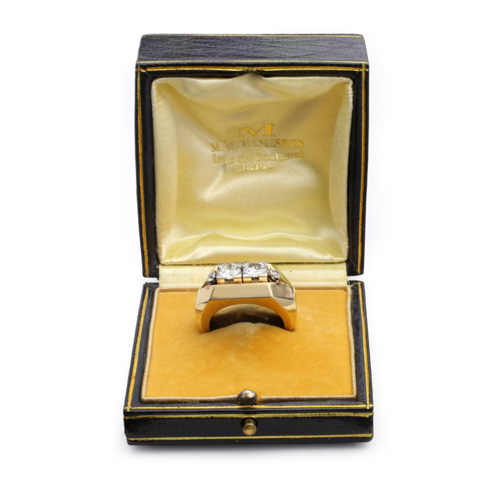 A Mauboussin Gold and Diamond Ring with three diamonds is displayed upright in an open vintage black and gold box featuring a yellow velvet interior and the Mauboussin brand name printed inside the lid.