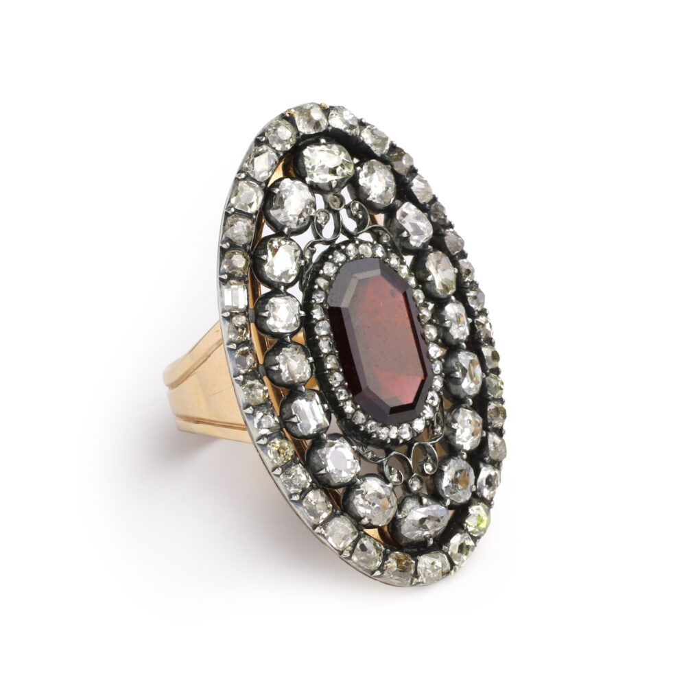 The Antique Garnet and Diamond Ring features an oval garnet centerpiece surrounded by rows of sparkling diamonds on a decorative gold band, showcased against a white background.