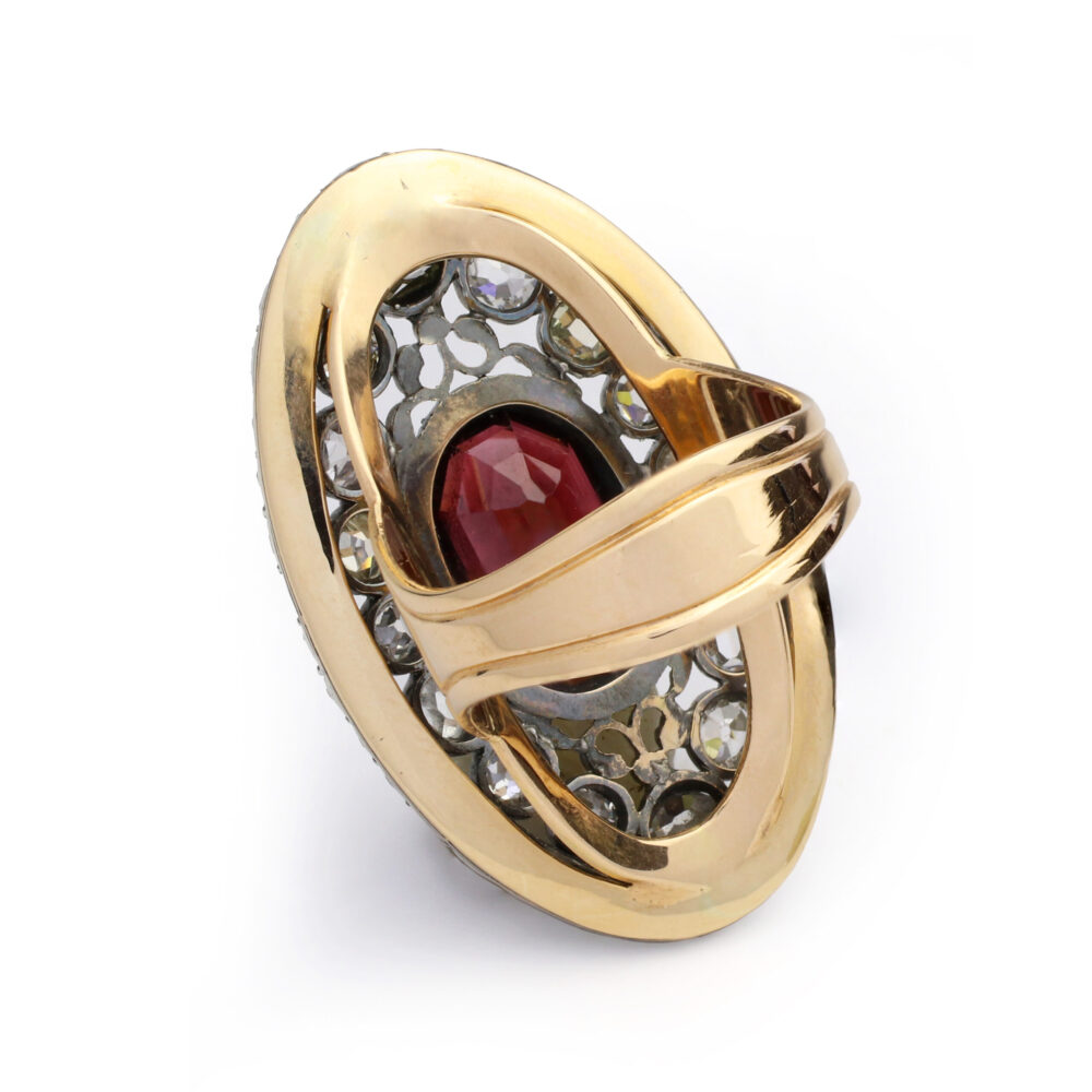 The Antique Garnet and Diamond Ring features an intricate lattice design with a large oval red center stone, small round clear accents, and a bold gold band crossing diagonally over the top.