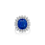 The Sapphire and Diamond Ring showcases timeless elegance and sophistication with its large oval Burmese blue sapphire center stone set in 18K white gold, encircled by a halo of small round white diamonds arranged in a floral-inspired design.