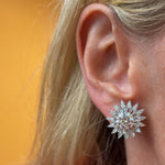 A close-up of a person's ear with long blonde hair, wearing the Radial Shield Diamond Ear Jackets in White Gold—large, round, starburst-shaped earrings featuring shield cut diamonds set in 18k white gold—against an orange background.