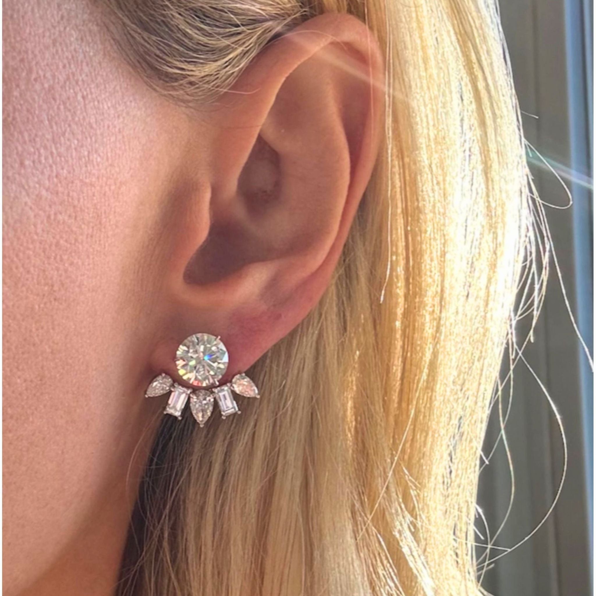 A close-up of a woman's ear showcases the Alternating Pear Shape and Emerald Cut Diamond Ear Jackets, featuring a dazzling emerald-cut diamond accented by five smaller, baguette-inspired stones arranged in an elegant fan shape reminiscent of modern ear jackets. Partially visible blonde hair and sunlight draw attention to the shimmering jewelry.