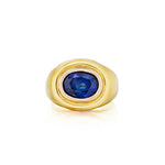 The 1970s Gold and Sapphire Ring features a striking, large round blue sapphire set at the center of a vintage gold mounting, elegantly presented against a white background.