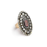 The Antique Garnet and Diamond Ring showcases a striking, cushion-cut garnet as its deep red centerpiece, elegantly encircled by two rows of sparkling diamonds in an oval setting that evokes the charm of a vintage treasure.