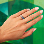 A hand with manicured nails displays the Sapphire and Diamond Ring—crafted in platinum with a geometric blue gemstone design—against a green abstract background.