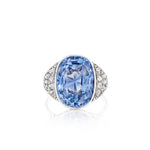 The 1930s Sapphire and Diamond Ring features a large oval Ceylon sapphire at its center, framed by clusters of small rose-cut diamonds on either side, all set in a sleek platinum or white gold band and displayed against a white background.