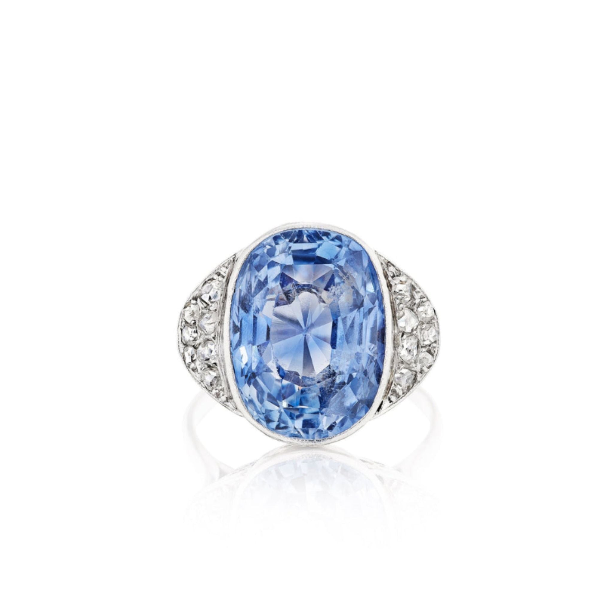 The 1930s Sapphire and Diamond Ring features a large oval Ceylon sapphire at its center, framed by clusters of small rose-cut diamonds on either side, all set in a sleek platinum or white gold band and displayed against a white background.