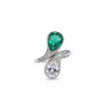 The Early 20th Century Emerald And Diamond Bypass Ring showcases a pear-shaped green Colombian emerald and a pear-shaped white diamond set at opposite ends of a platinum bypass band, which is further adorned with small diamonds.