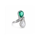 The Early 20th Century Emerald And Diamond Bypass Ring is crafted in platinum and showcases a pear-shaped Colombian emerald paired with a pear-shaped clear diamond, each encircled by delicate white accent stones in an elegant bypass design.