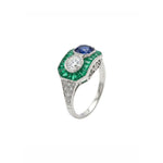 The Sapphire, Emerald and Diamond Ring is a vintage platinum piece showcasing a round blue sapphire and a round white diamond, accented with green emerald baguettes and small old European-cut diamonds on a white metal band.