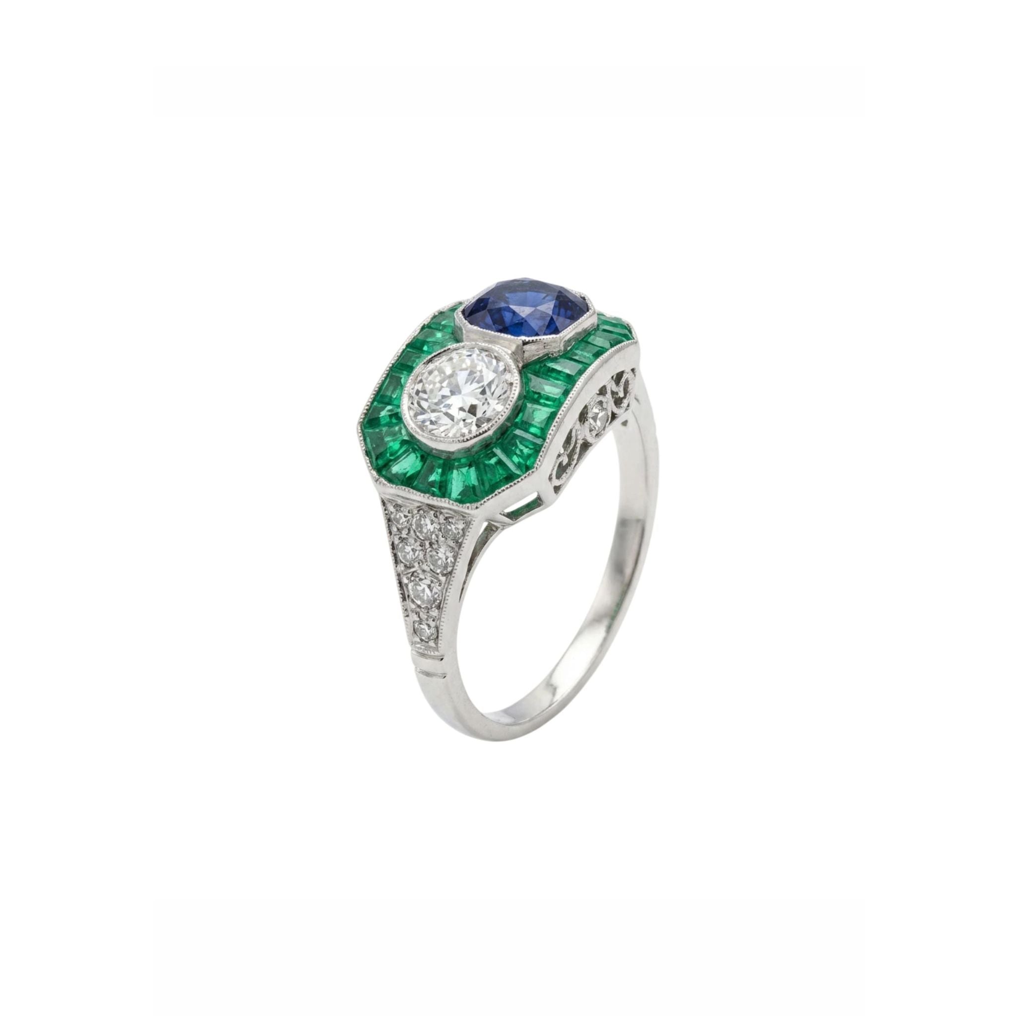 The Sapphire, Emerald and Diamond Ring is a vintage platinum piece showcasing a round blue sapphire and a round white diamond, accented with green emerald baguettes and small old European-cut diamonds on a white metal band.