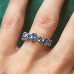 A hand wearing the Sapphire Mixed Shape Cluster Ring rests on green fabric; the ring features sapphires of varied shapes set in silver.
