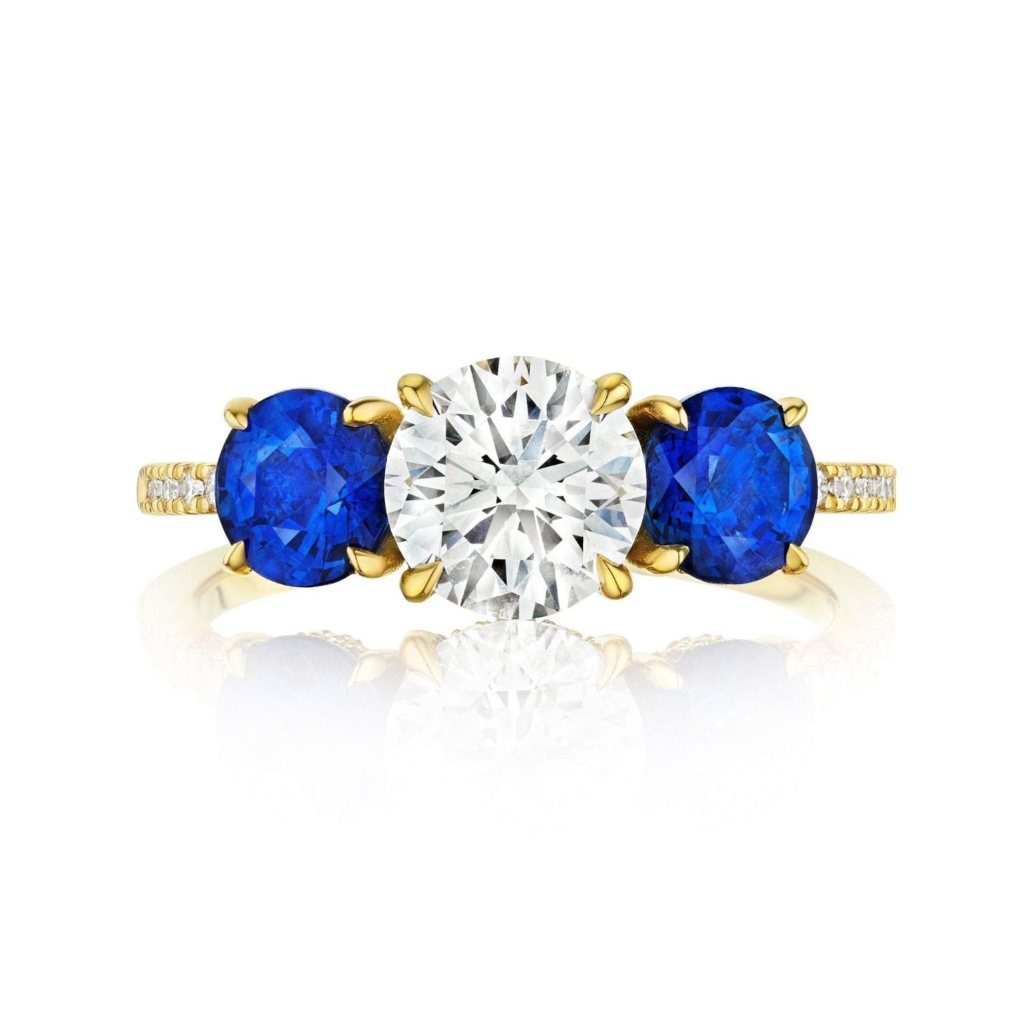 The Round Diamond Engagement Ring with Round Sapphire Side Stones features a large round brilliant diamond center, two round blue sapphire sides, and small accent diamonds along the 18K yellow gold band.