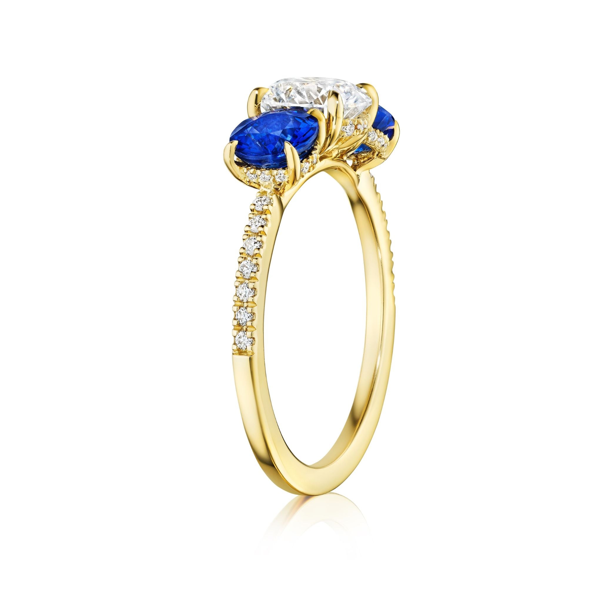 The Round Diamond Engagement Ring with Round Sapphire Side Stones features an 18K yellow gold band set with small diamonds, a large round brilliant diamond center, and two round blue sapphires, displayed on a white background.