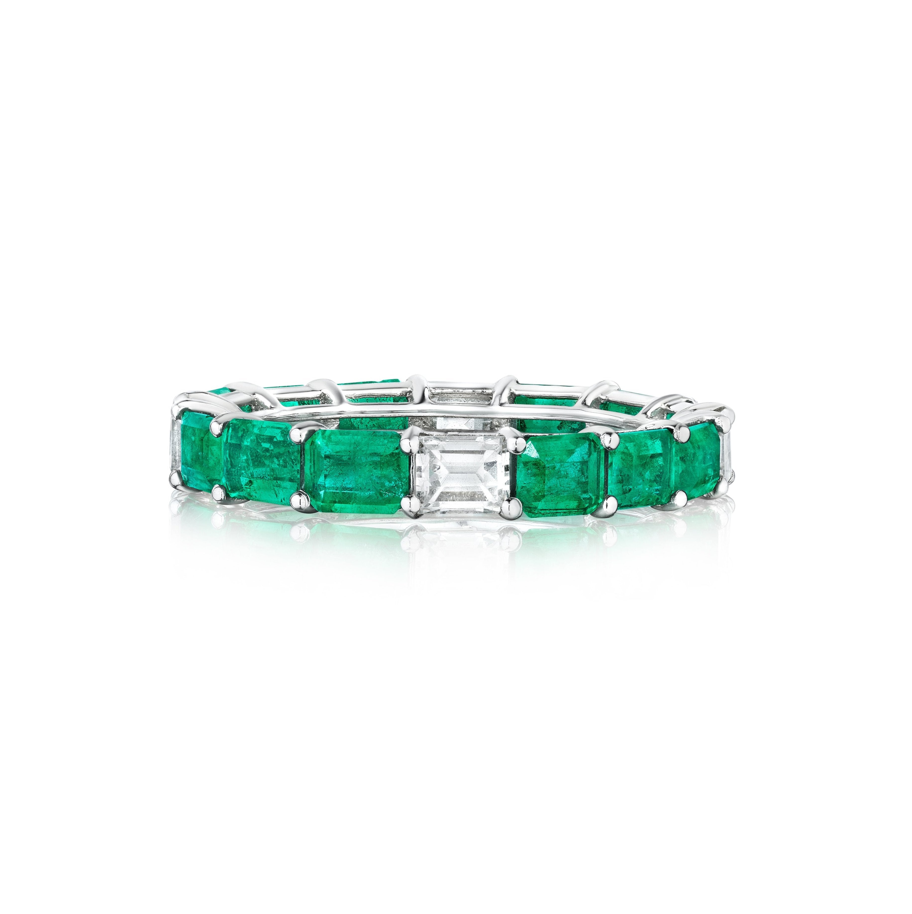 The East-West Emerald Cut Emerald and Diamond Eternity Band in White Gold showcases alternating rectangular emeralds and baguette-cut diamonds in a prong setting, displayed on a white background.