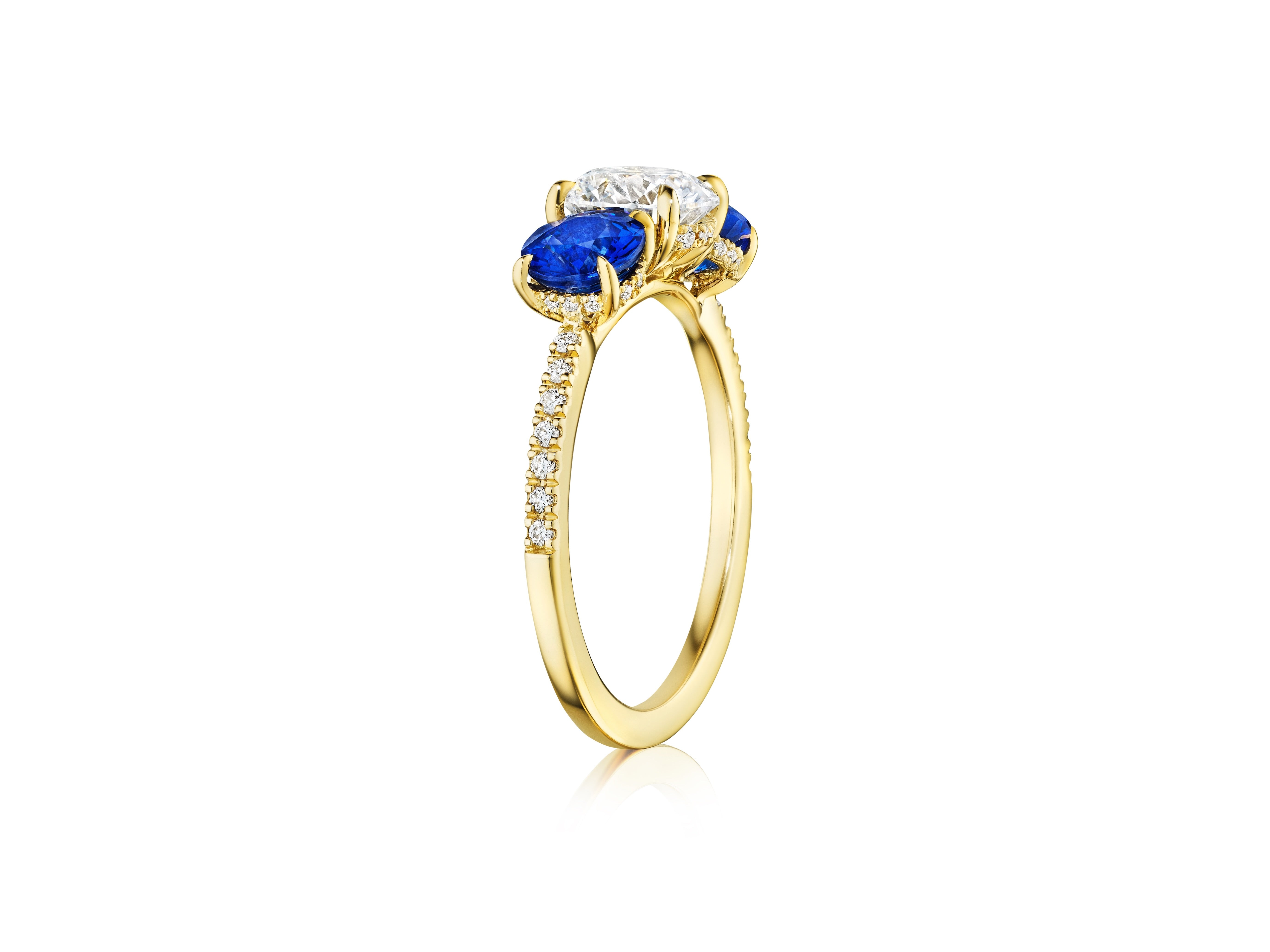 The Round Diamond Engagement Ring with Round Sapphire Side Stones features a central round diamond, two blue sapphires, and smaller diamonds on an 18K yellow gold band, shown upright against a white background.