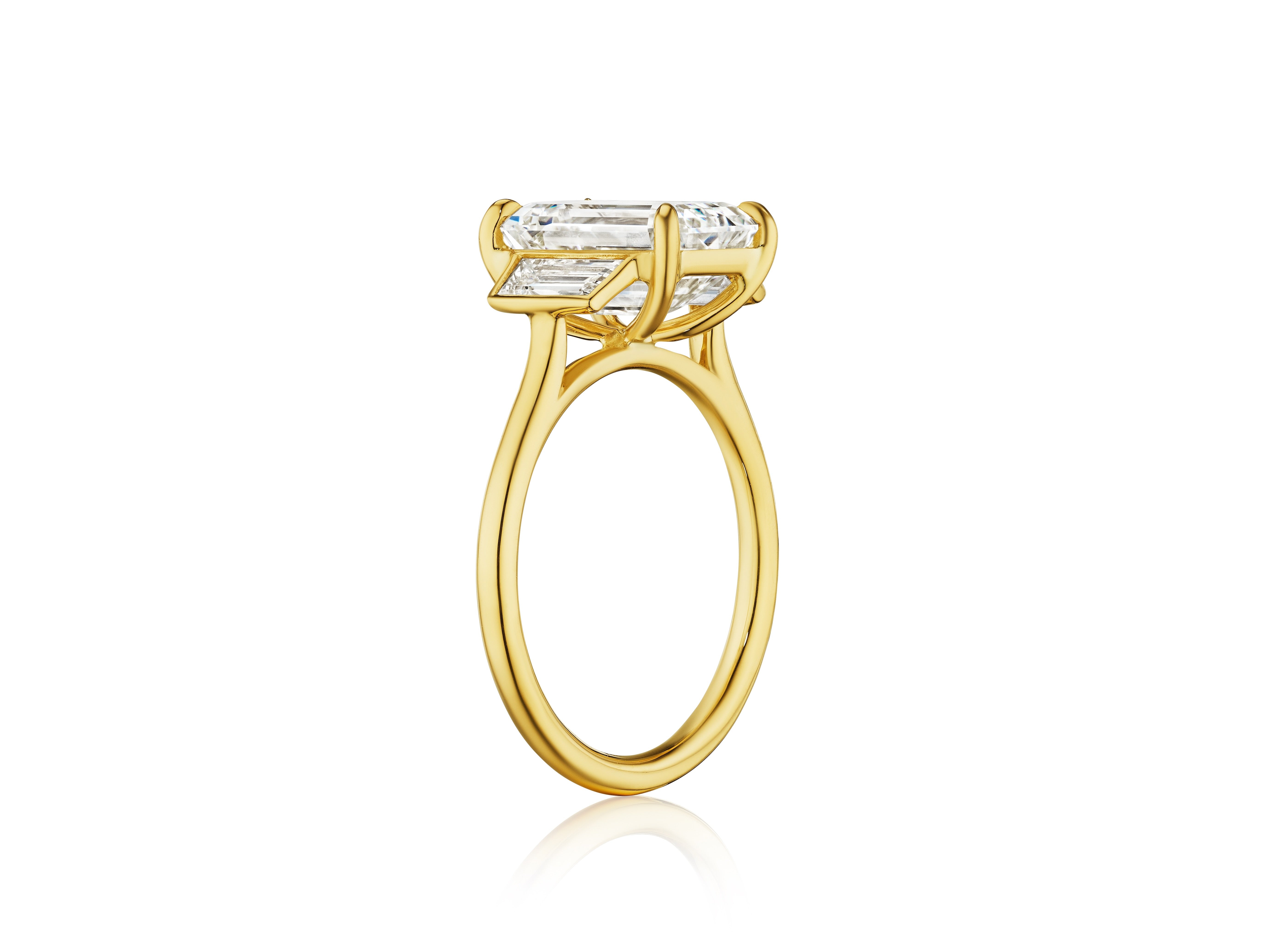 The Emerald Cut Diamond Engagement Ring with Trapezoid Side Stones in yellow gold is shown upright, with its emerald cut diamond in a four-prong setting, against a plain white background.