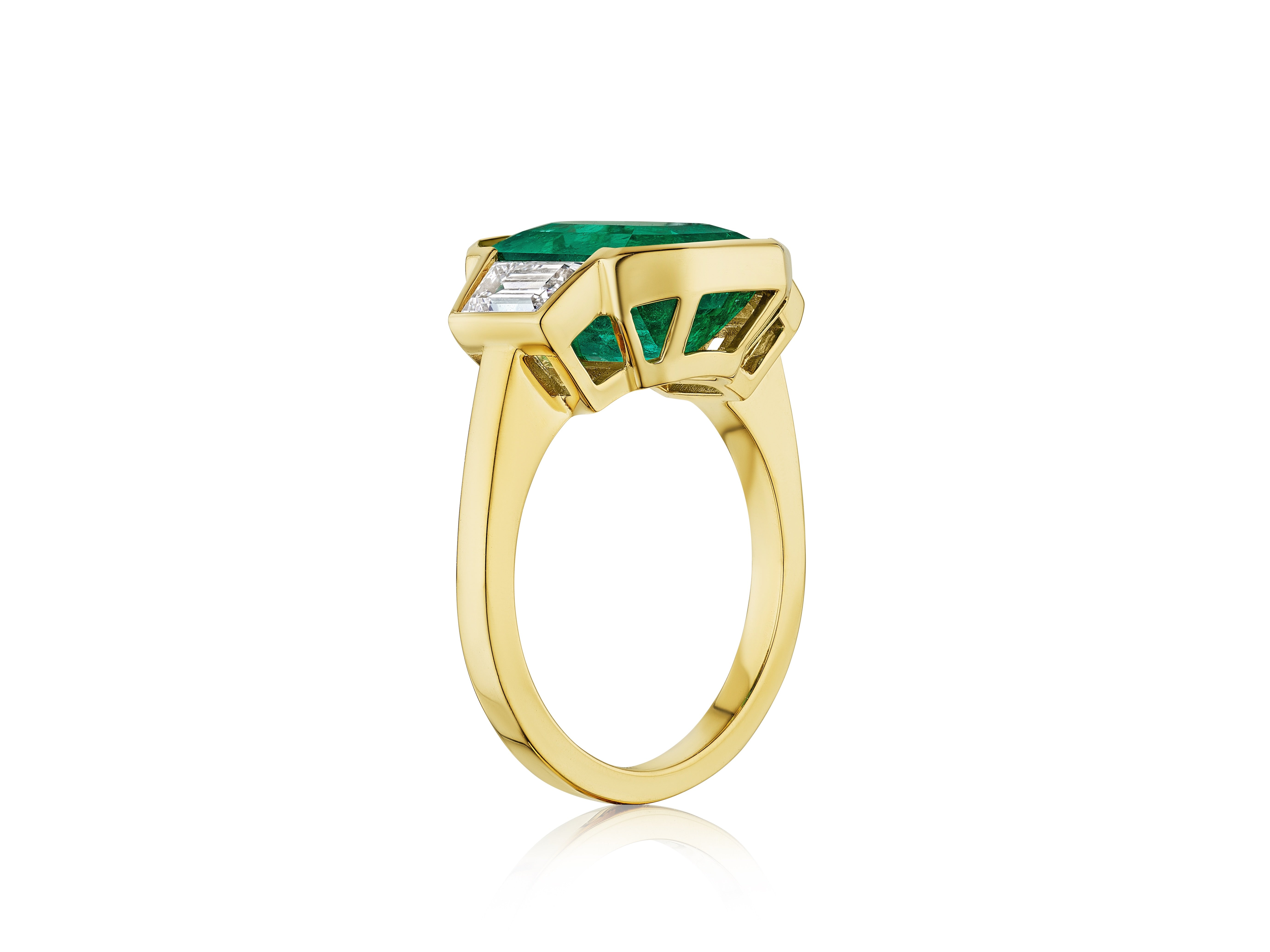 The Half Bezel Set Emerald Cut Emerald Engagement Ring features a raised 18k yellow gold setting with trapezoid diamond side stones, displayed on a white background.