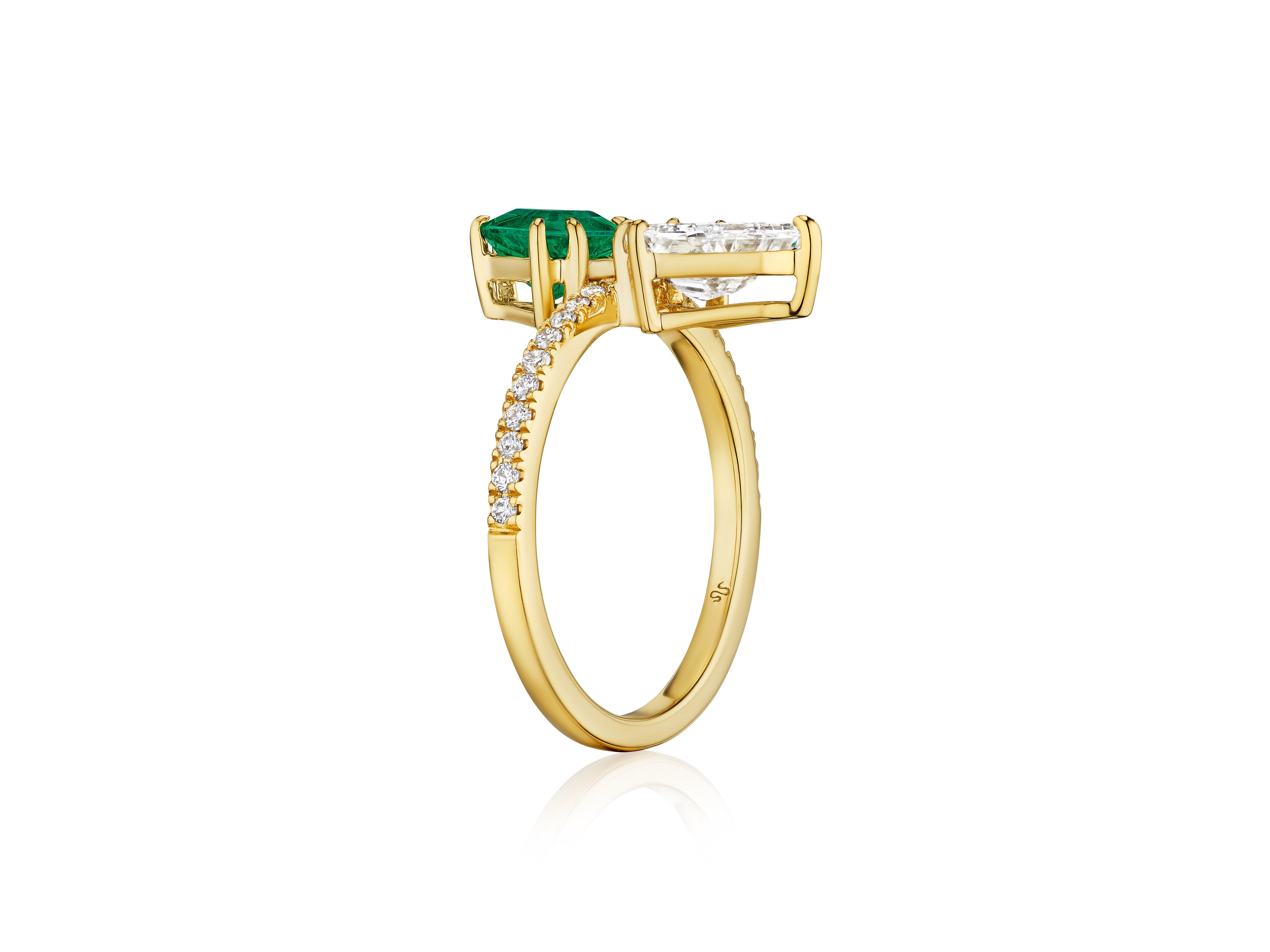 The Emerald and Diamond Double Shield Ring in Yellow Gold features a modern design with a rectangular green gemstone and clear gemstone set side by side, partially encrusted with small diamonds for added brilliance.