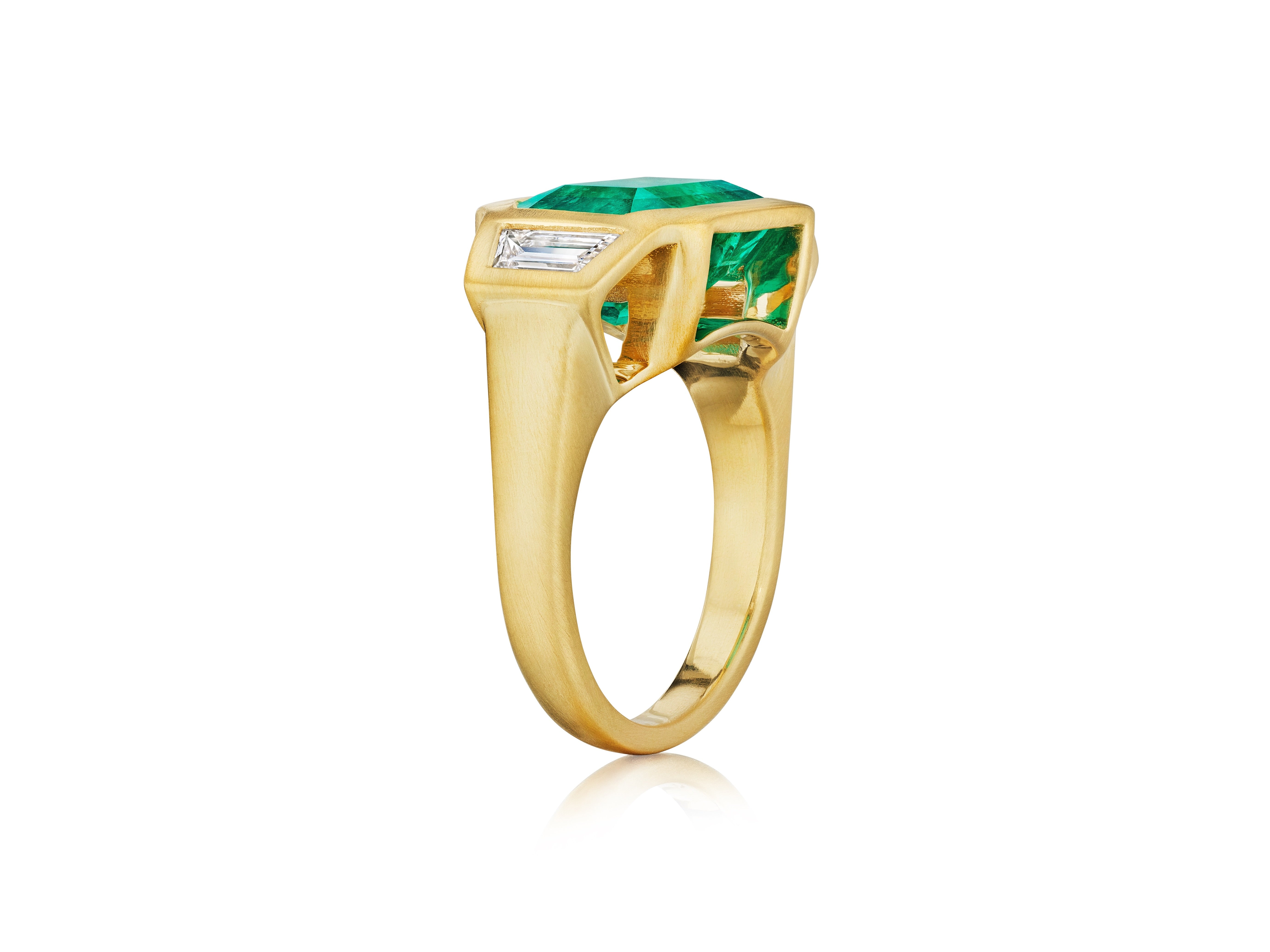 The Bezel Set East-West Emerald Cut Emerald Three Stone Ring features trapezoid diamond side stones in matte yellow gold, photographed on a white background.