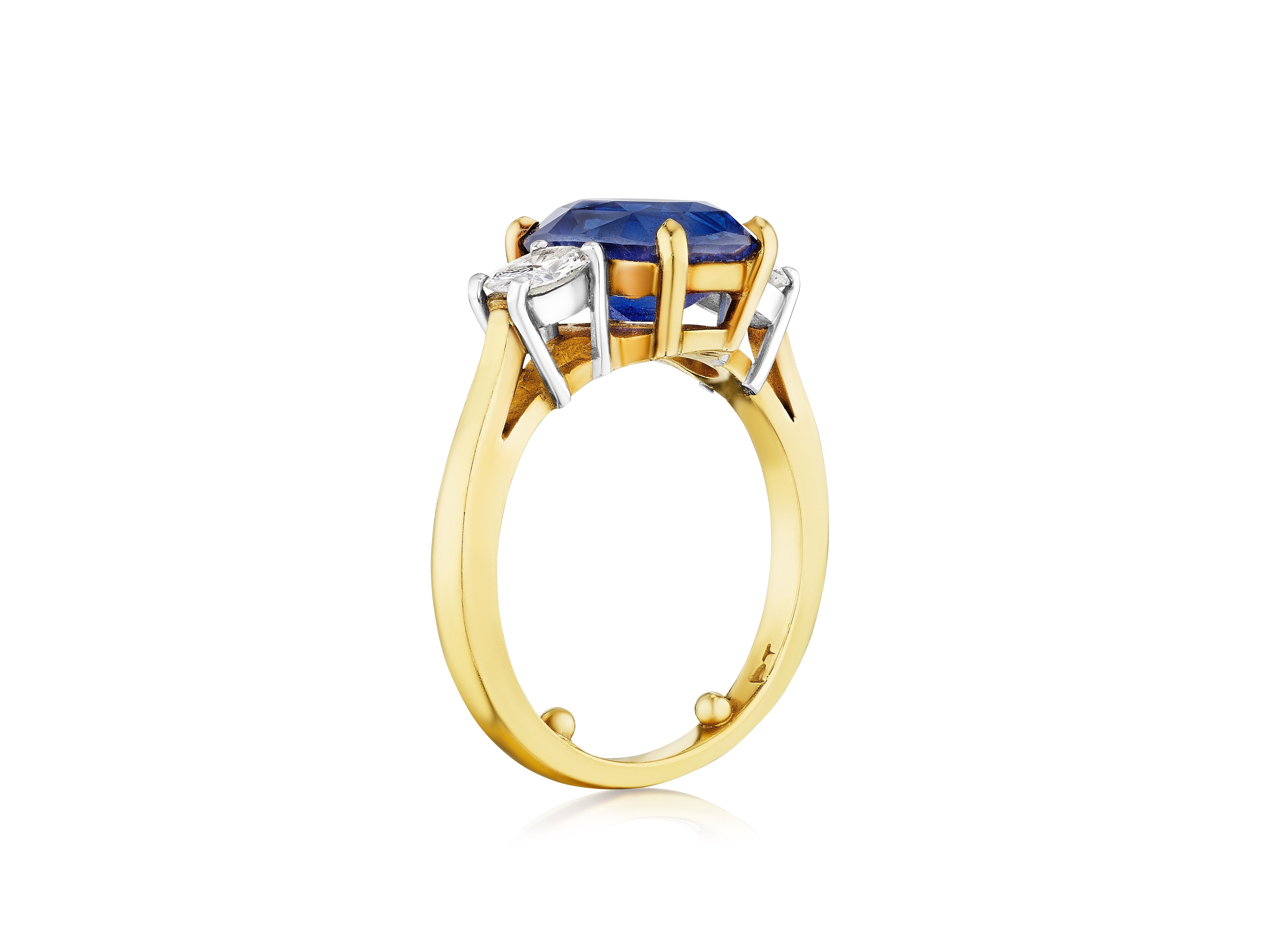 The Cushion Cut Blue Sapphire Engagement Ring in Yellow Gold features a prong-set blue sapphire and oval diamond side stones, displayed against a white background.