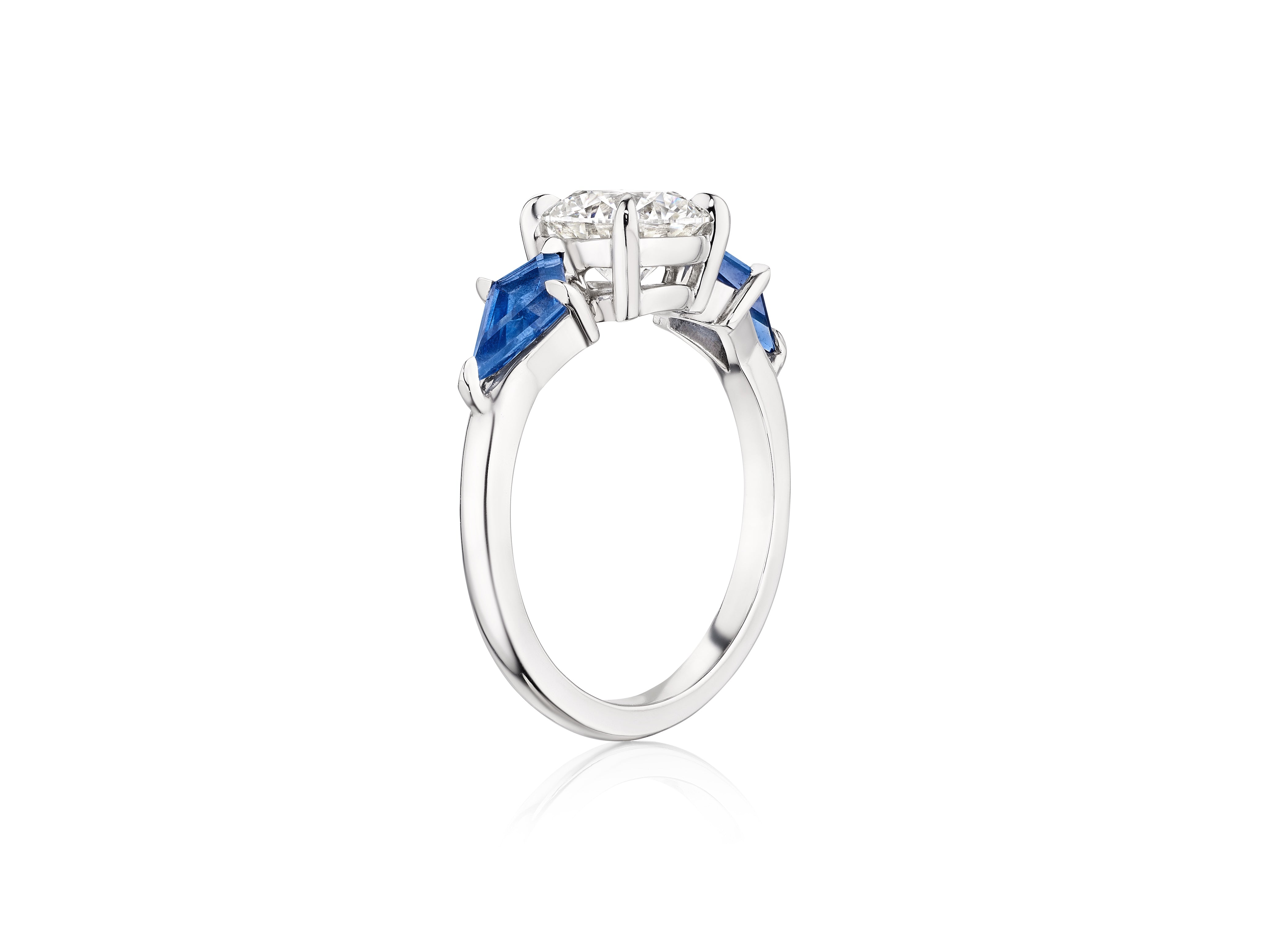 The Bespoke Round Diamond Engagement Ring in White Gold features a central round diamond and kite-shaped blue sapphire side stones, shown from a side angle on a white background.