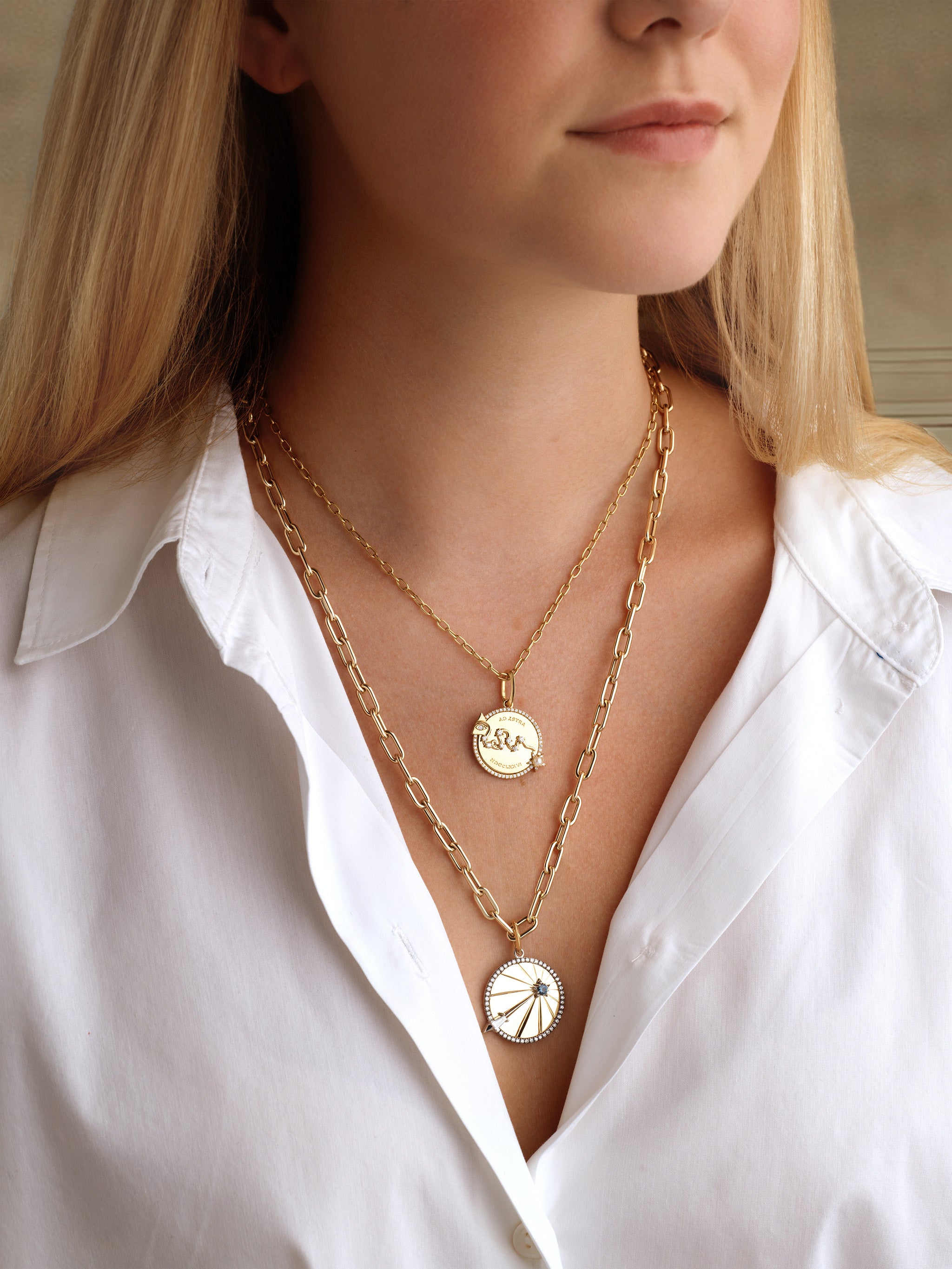 A woman with long blonde hair wears a white button-up shirt and two layered gold necklaces: one features the America250 Ad Astra Pavé Diamond and Pearl Medallion Pendant in yellow gold; the other showcases a sunburst design.