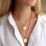 A woman in a white button-down shirt models two America250 Pavé Star Medallion Pendants in yellow gold, each featuring intricate details. The neutral background accentuates the handcrafted New York jewelry.