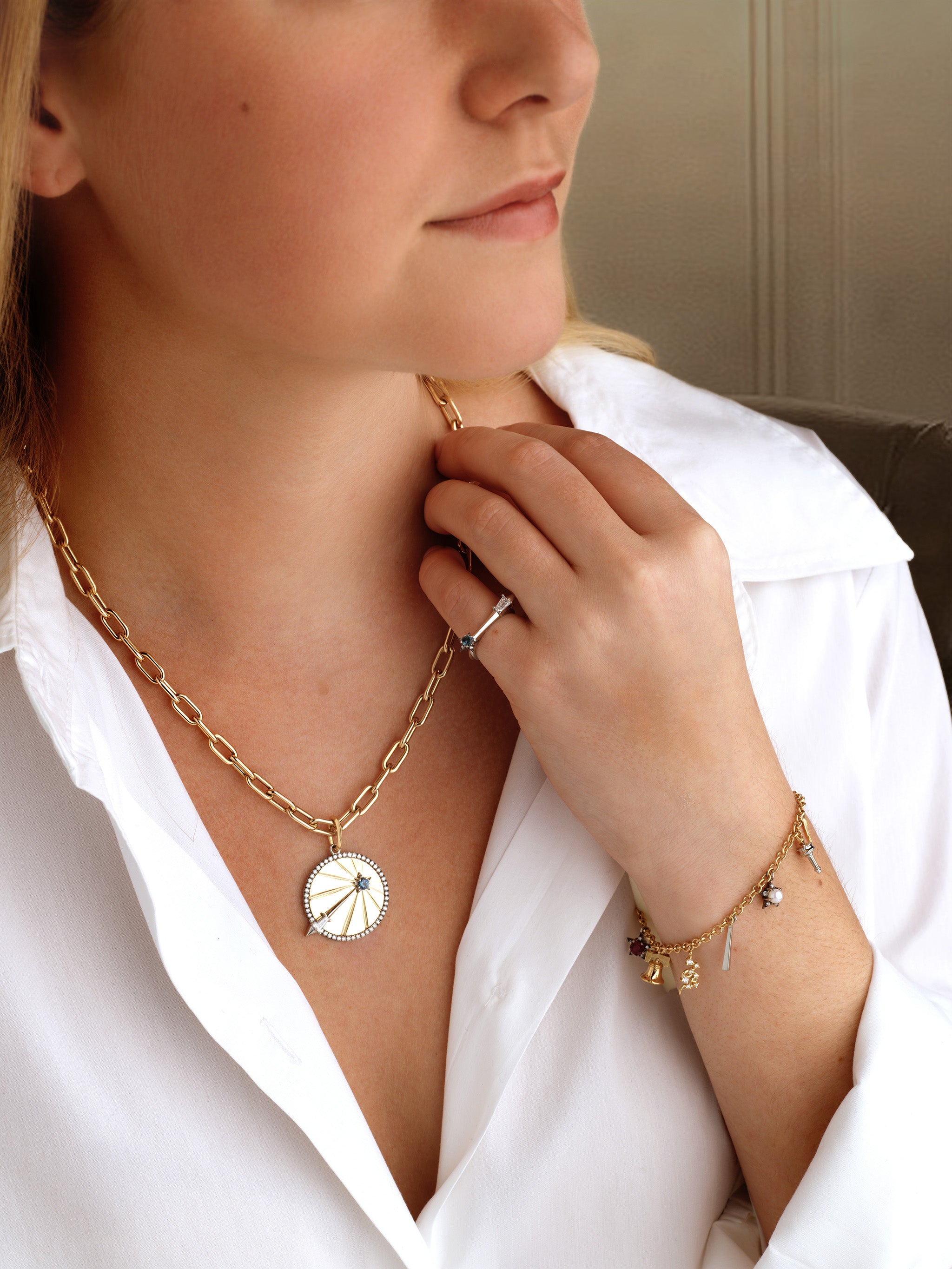 A woman in a white blouse showcases gold jewelry from the America250 capsule collection: an 18k yellow gold chain necklace with a round pendant, a ring, and the America250 Ruby, Pearl, Sapphire, and Diamond Charm Bracelet. She touches her necklace gently.