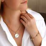 A woman in a white blouse models handcrafted New York jewelry, featuring the America250 Pavé Star Medallion Pendant in Yellow Gold on a chunky chain necklace, paired with a charm bracelet and ring. She gazes slightly to the side with a relaxed look.