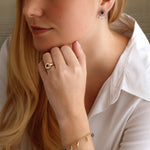 A woman with long blonde hair and a white collared shirt showcases jewelry, including the America250 Star Signet Ring in Yellow Gold and Platinum, a charm bracelet, and colorful stud earrings. She looks to the side with her hand near her face.