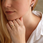 A woman with long blonde hair in a white shirt rests her chin on her hand, wearing the America250 Star Signet Ring in Yellow Gold and Platinum and silver cluster earrings. Her calm, thoughtful expression evokes quiet elegance.