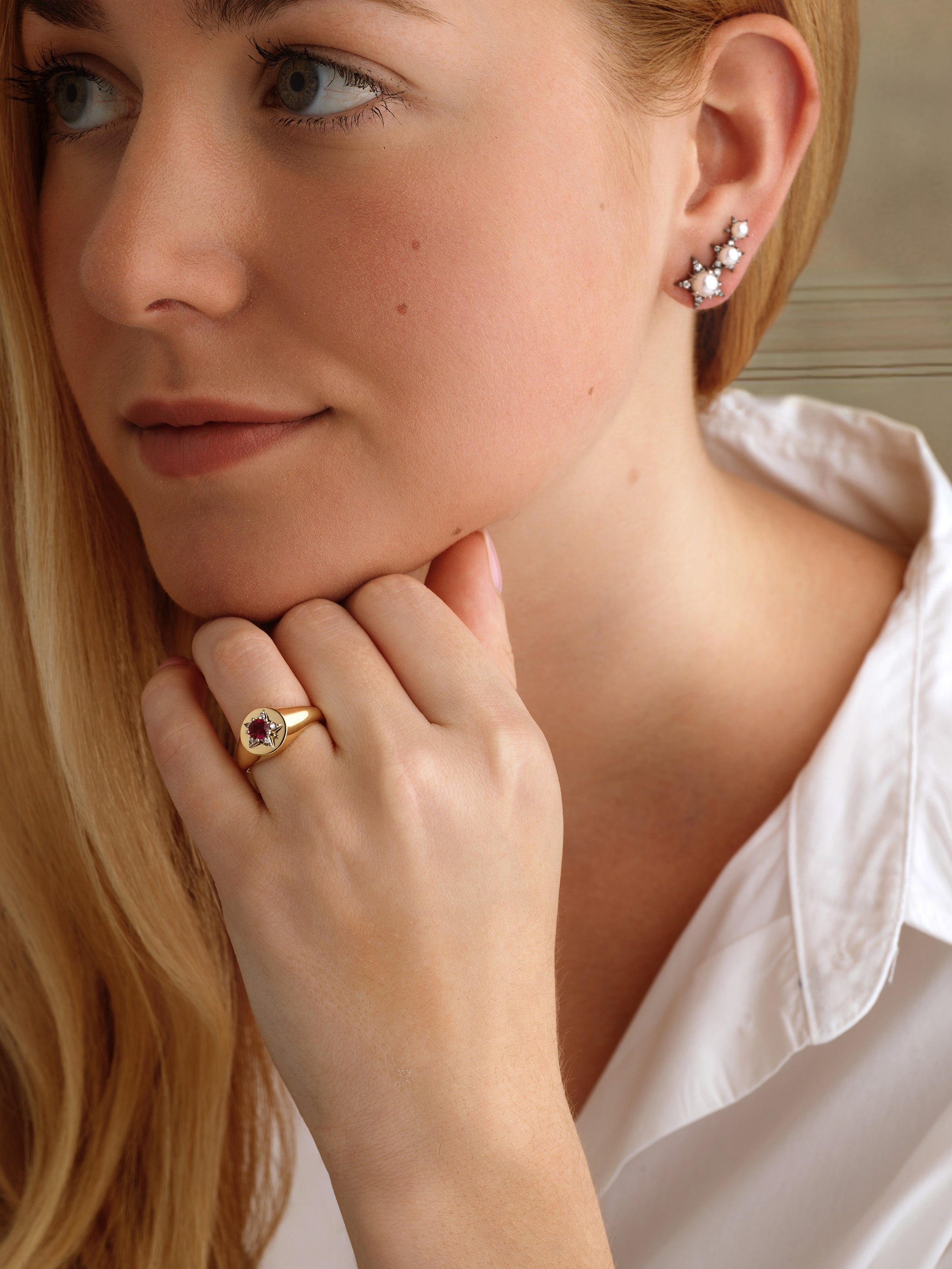 A woman with long blonde hair wears a white shirt, a gold gemstone ring, and America250 Trio Star Crawler Earrings in Black Rhodium—limited-edition jewelry handcrafted in NYC. She rests her chin on her hand and gazes calmly to the side.