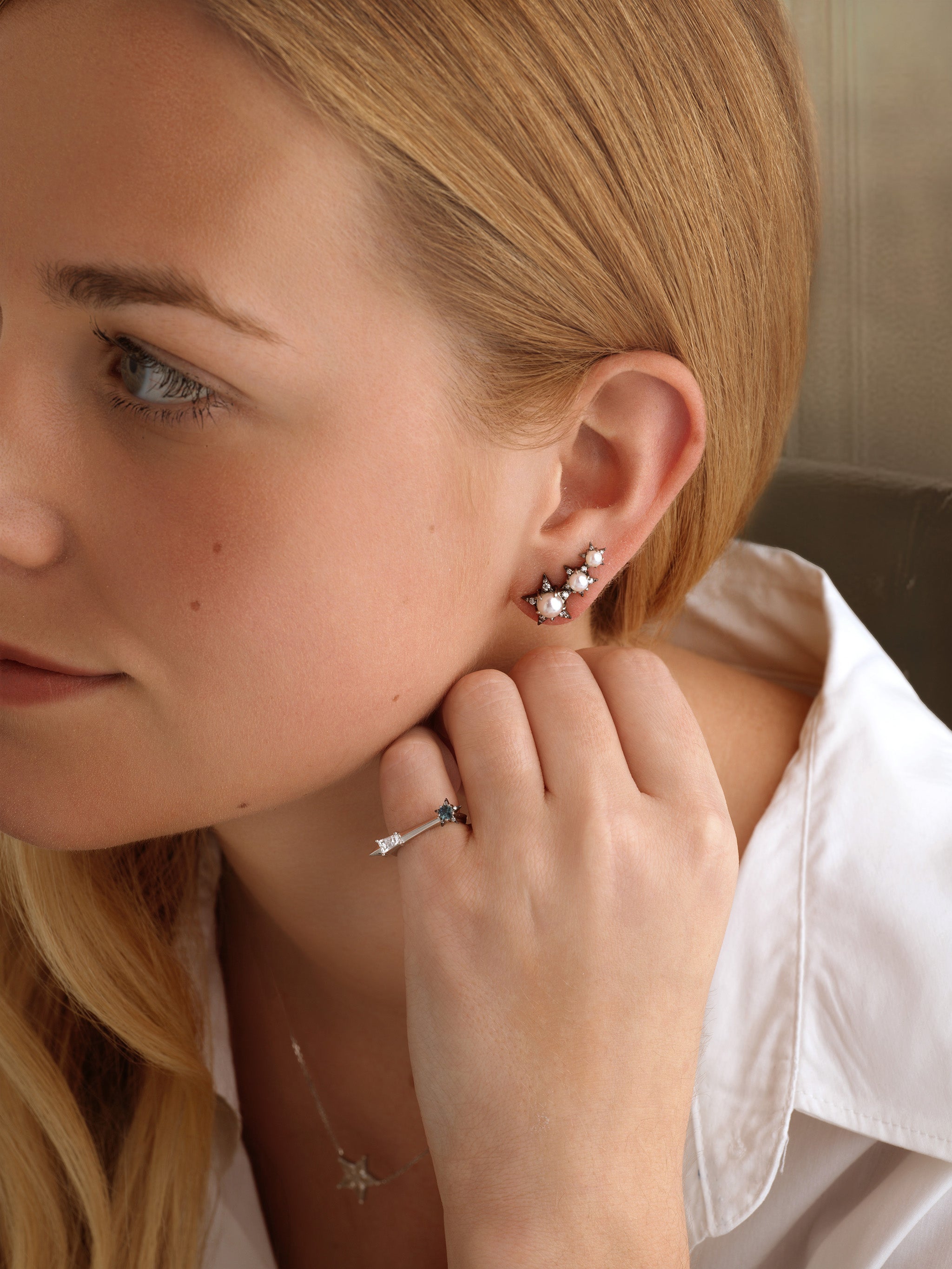 A woman with long blonde hair, in a white shirt and silver floral earrings, rests her hand near her ear wearing the America250 Star and Baguette Sapphire and Diamond Ring in Platinum, along with a delicate star-shaped necklace.
