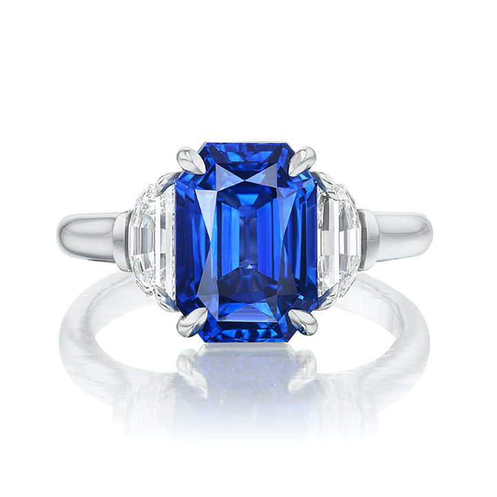 The Emerald Cut Sapphire Engagement Ring features a striking emerald-cut blue sapphire center stone with half moon diamond side stones, set in a polished platinum band for a stunning and elegant look.