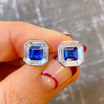 A hand with red nail art holds Mosaic Studs in White Gold, each featuring an Asscher cut sapphire center and surrounded by baguette diamonds, against a blurred background.