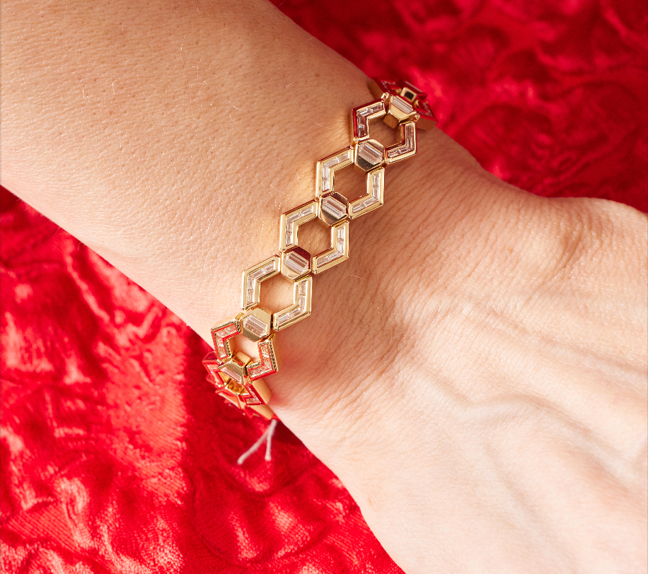 The Chrysler Hexagon Bangle with Baguette Diamonds, inspired by Art Deco style and featuring hexagonal links, is shown on a wrist above a red textured fabric background.
