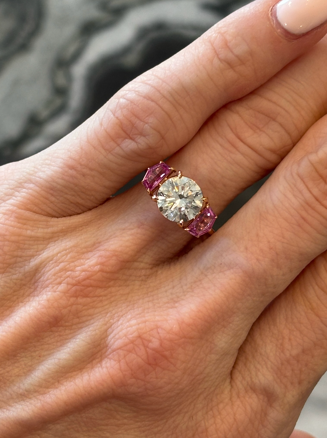 A hand with light skin tone wears the Round Brilliant Diamond Engagement Ring with Epaulette Pink Sapphire Side Stones, featuring a large round diamond and rectangular pink sapphires, with neatly manicured nails visible.
