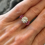 A hand with light skin tone wears the Round Brilliant Diamond Engagement Ring with Epaulette Pink Sapphire Side Stones, featuring a large round diamond and rectangular pink sapphires, with neatly manicured nails visible.