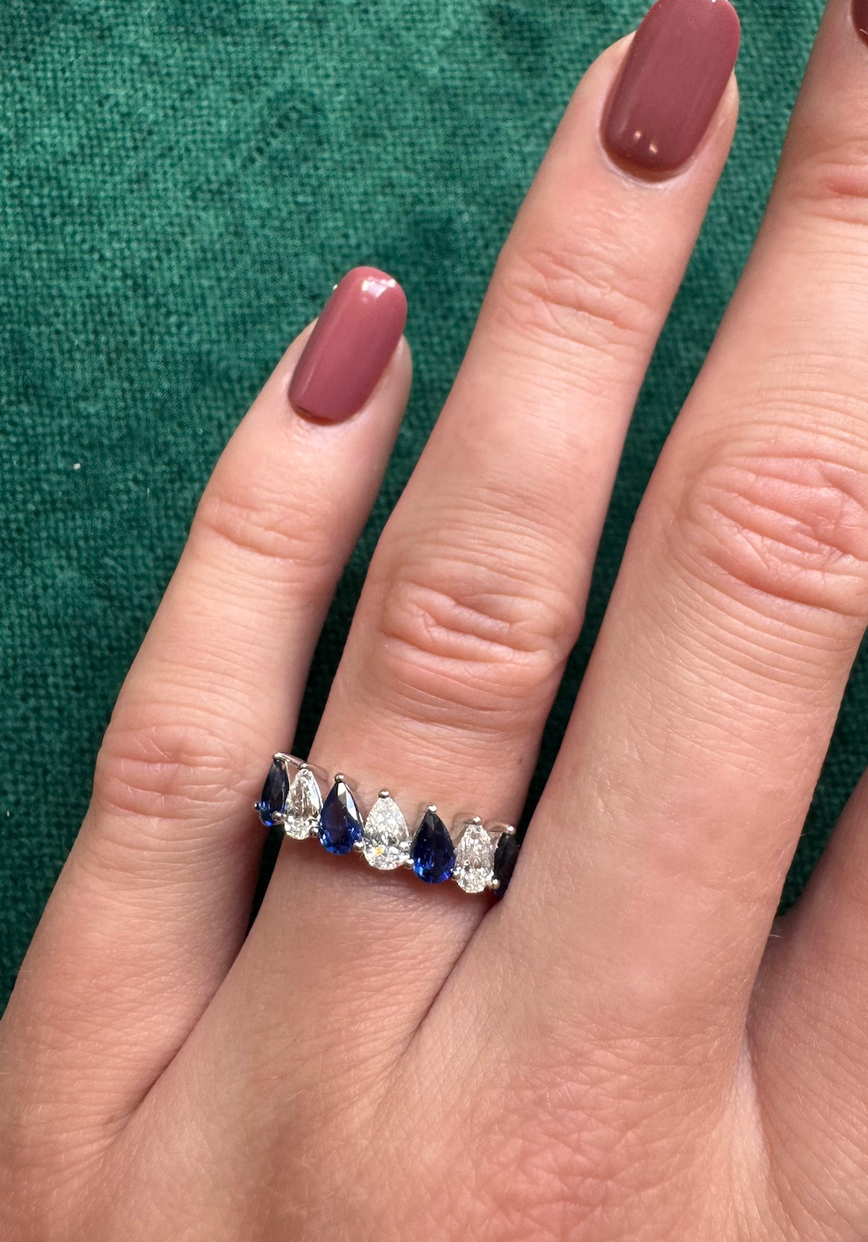 A hand with mauve-painted nails wears the Diagonal Pear Eternity Band with Alternating Diamonds and Pears—silver, set with blue and clear pear-shaped stones—against a green fabric background.