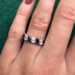 A hand with mauve-painted nails wears the Diagonal Pear Eternity Band with Alternating Diamonds and Pears—silver, set with blue and clear pear-shaped stones—against a green fabric background.