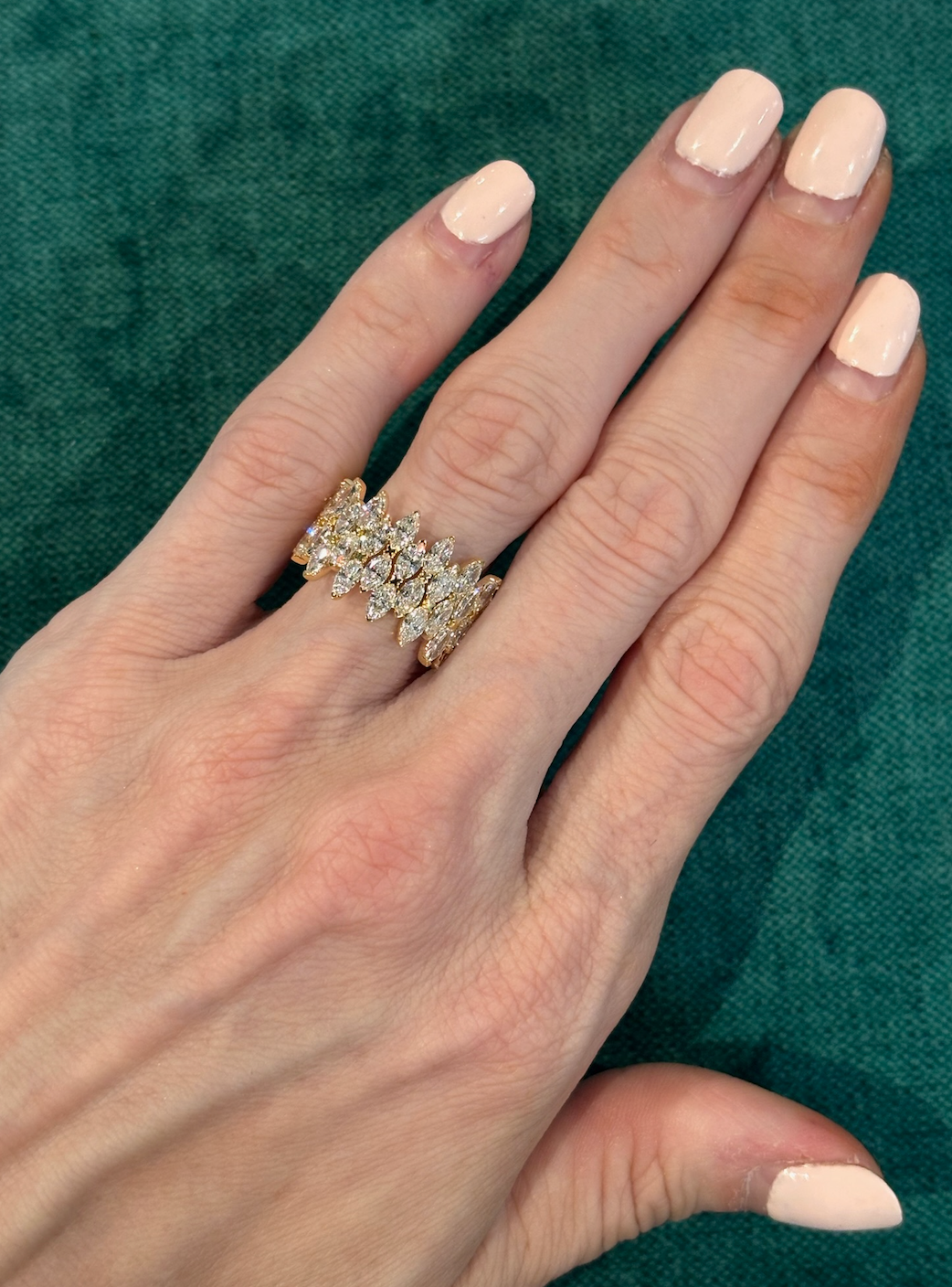 A hand with pale pink polished nails wears the Multi Tier Marquise Diamond Eternity Band, an ornate 18k gold ring featuring marquise-cut diamonds in a crisscross, vine-like multi-tier design, set against a textured green background.
