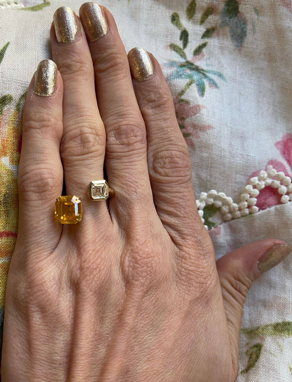 A hand with gold glitter nail polish displays the Yellow Sapphire and Diamond Toi et Moi Ring, featuring striking rectangular gemstones, against floral fabric with pearl accents.